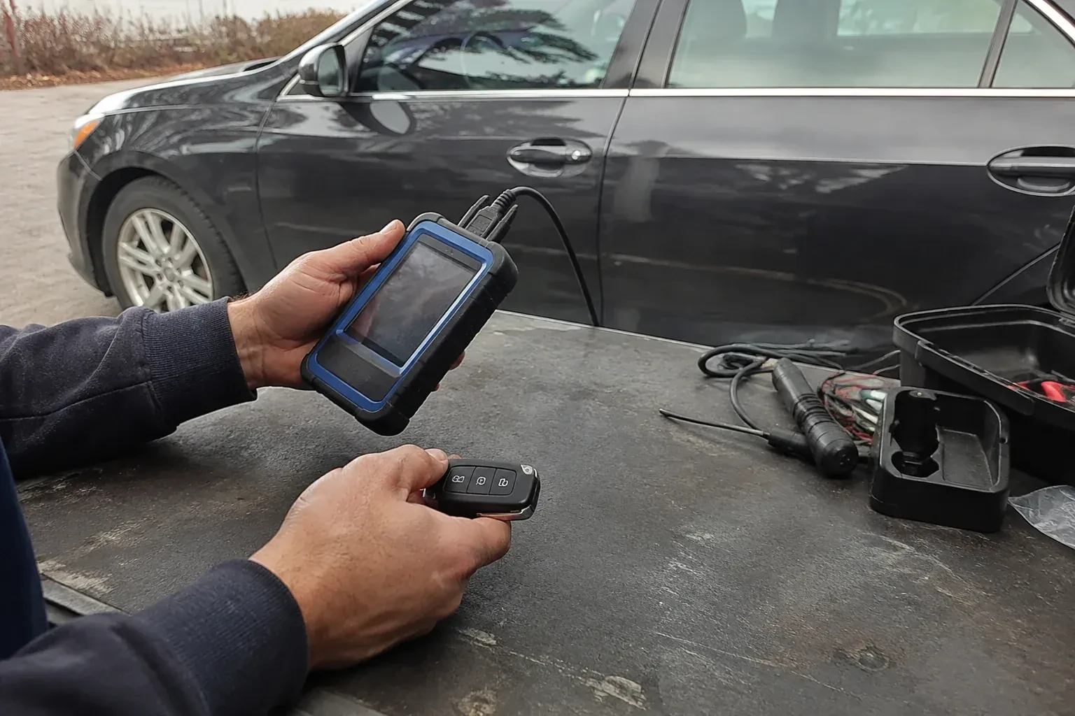 Hands holding a blue-edged electronic device and car-key fob at a worktable beside a parked car. — Transponder Key Programming, Oakville