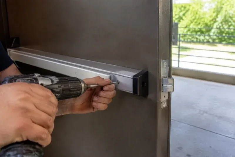 Hands using a power driver at the end of a horizontal push bar on an open metal door. — Panic & Exit Hardware, Oakville